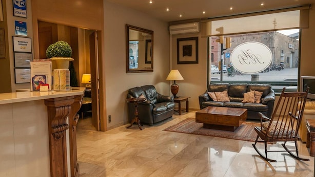 A modern hotel lobby with a reception desk, leather sofas, a wooden coffee table, and a large window offering a view of the street. - Ramón Berenguer IV (Lleida)