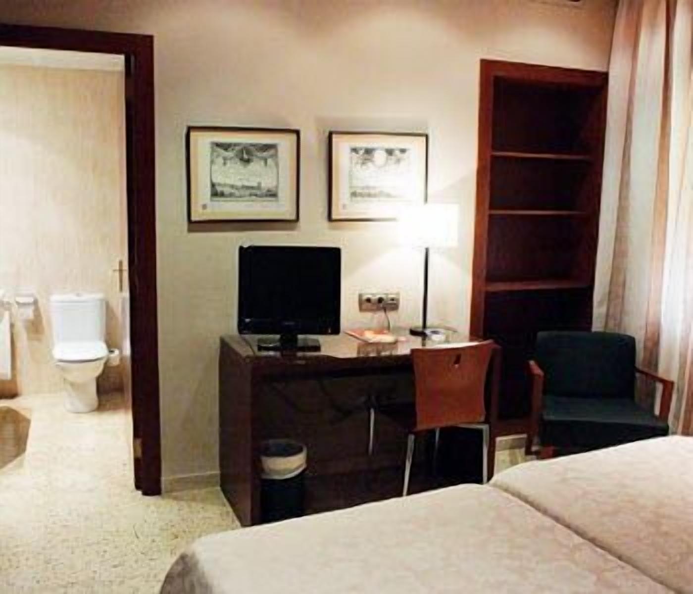 A hotel room with a bed, a desk with a computer, a chair, a TV, and a bookshelf.