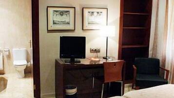 A hotel room with a bed, a desk with a computer, a chair, a TV, and a bookshelf.