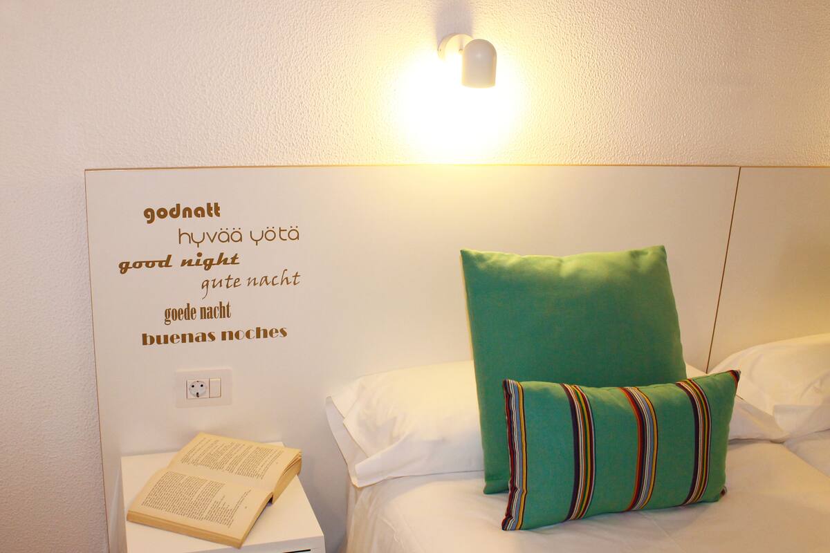 A neatly made bed with a green pillow and striped pillowcase, a bedside table with an open book, and a wall with multilingual 'good night' text.