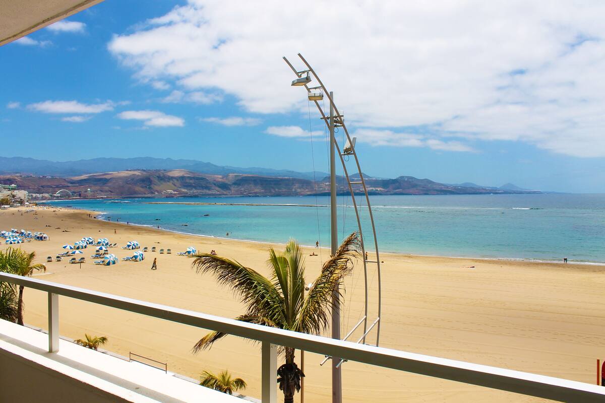 A beach with a clear view of the ocean and distant mountains. There are umbrellas and people on the beach.