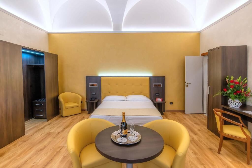 A hotel room with a bed, a bedside table with a bottle and glasses, a chair, a small table with a tray, and a wardrobe.
