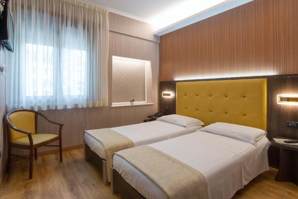 Ein Hotelzimmer mit zwei Betten, einem Sessel, einem Schreibtisch und einem Fenster mit Vorhängen.