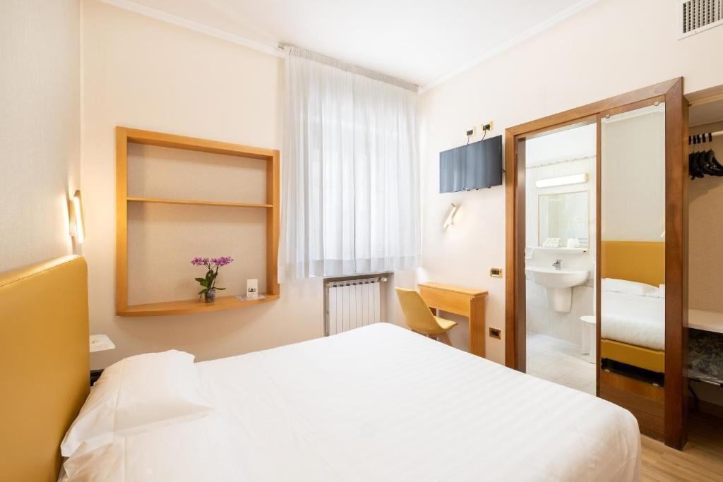 A hotel room with a bed, a desk, a chair, a mirror, a shelf with a vase, and a bathroom visible through an open door.
