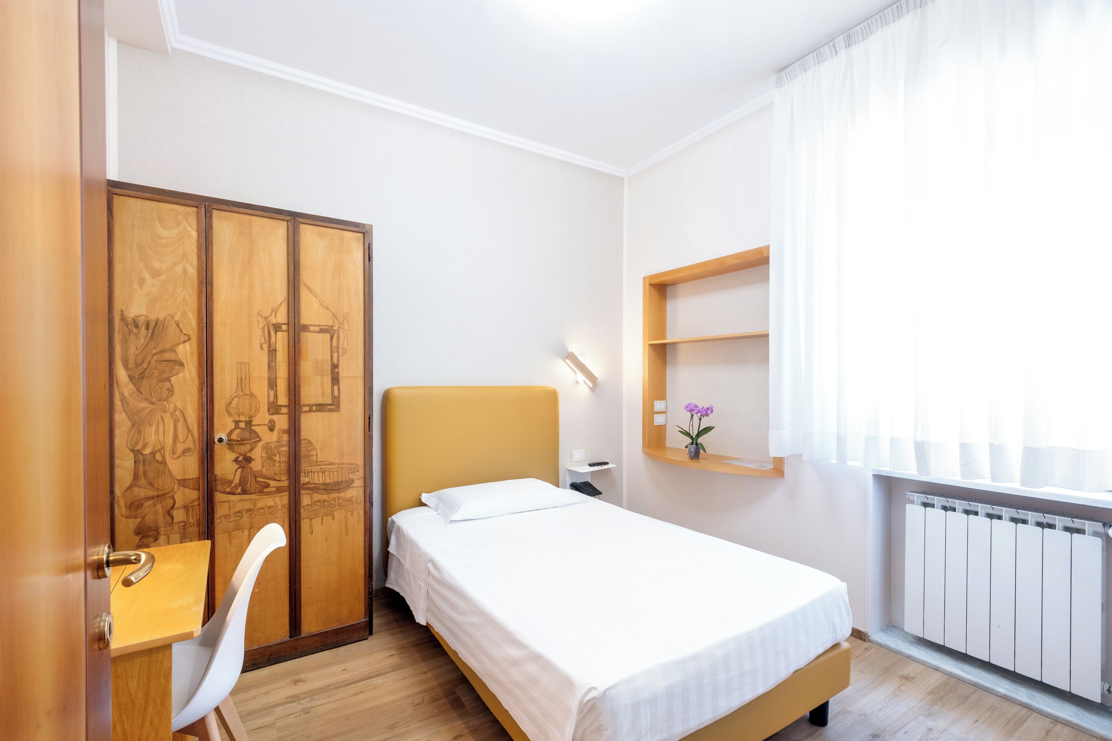 A bedroom with a bed, a desk, a chair, a wardrobe, a shelf with a vase, and a radiator.