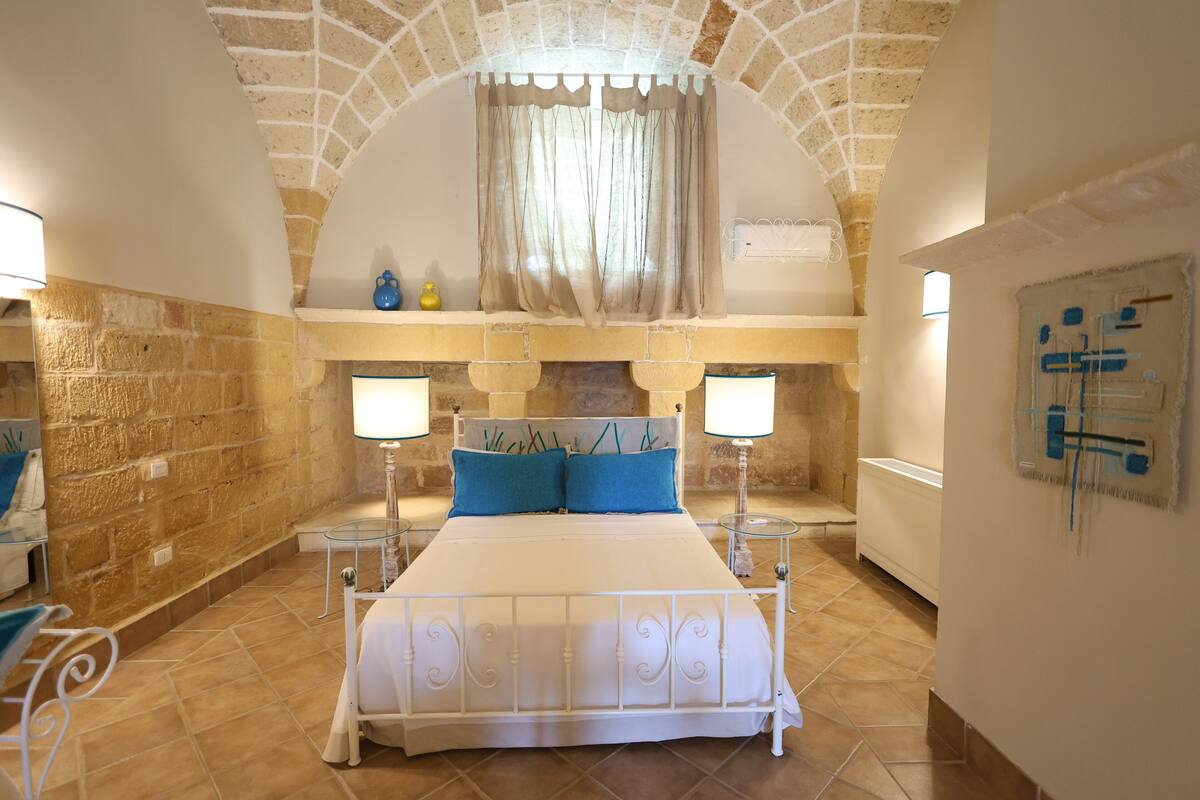 A bedroom with a stone wall, a bed with white metal frame, two blue pillows, and two bedside lamps.