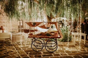 A rustic stone floor with a cart and wicker chairs, decorated with hanging lights and greenery.