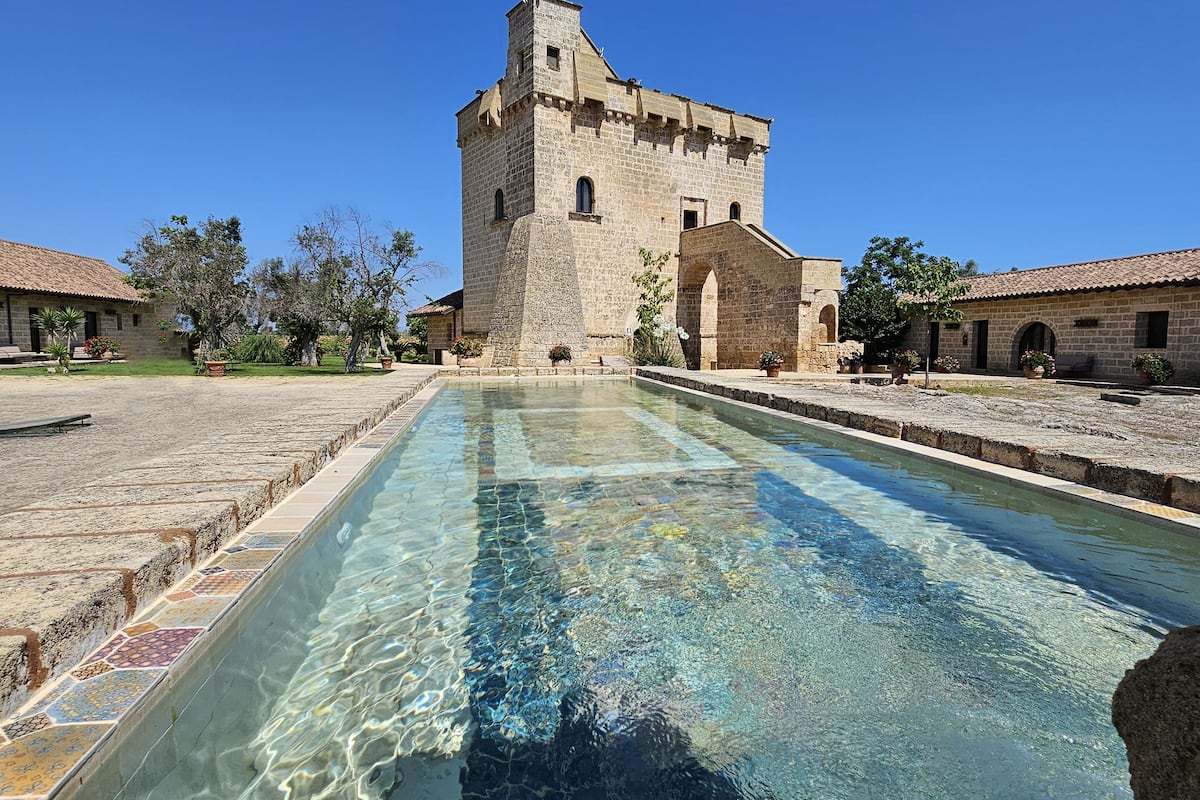 A clear pool with a stone border, a stone building with a tower, and a clear blue sky.
