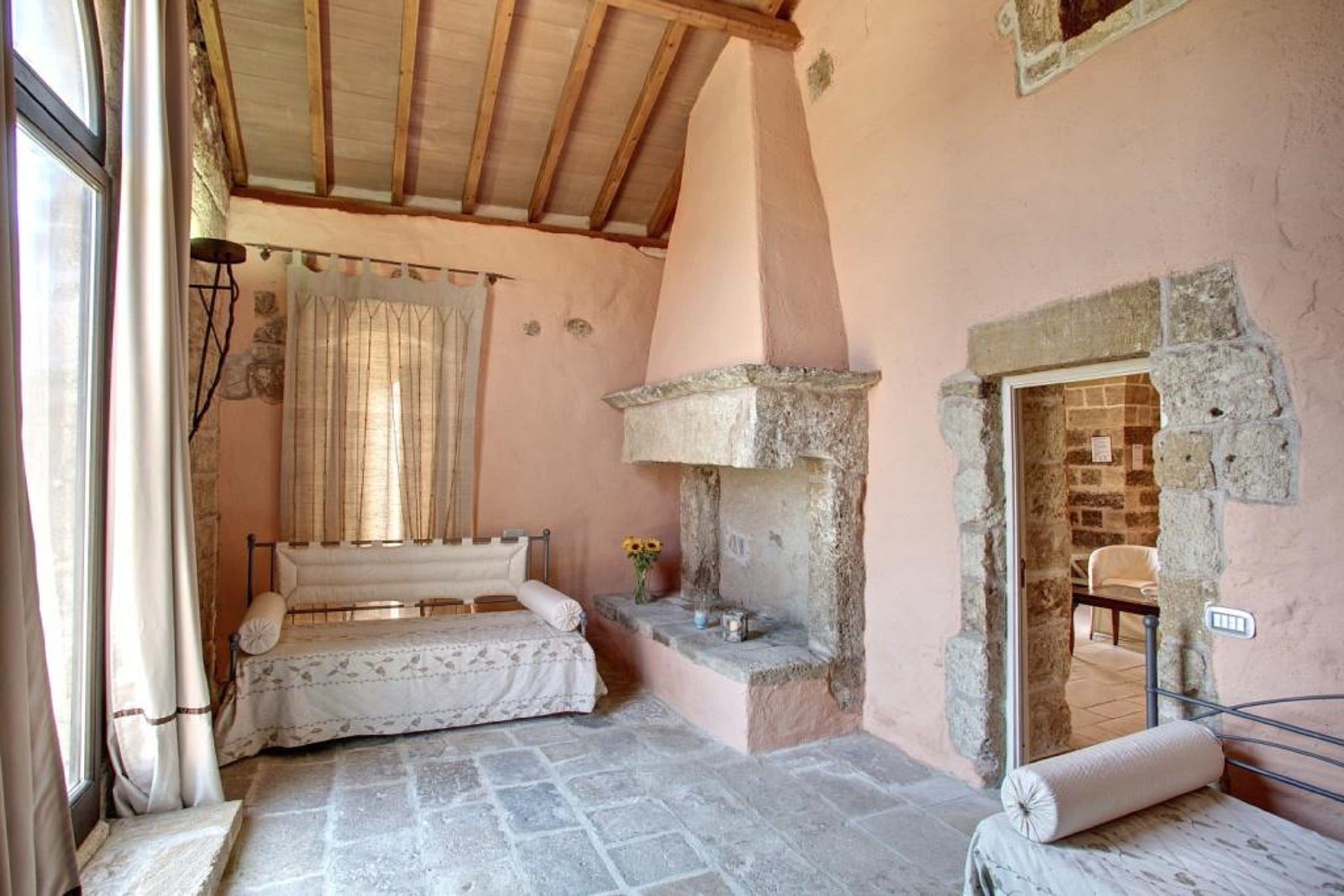 A rustic bedroom with a stone fireplace, a bed with a floral bedspread, and a window with curtains.