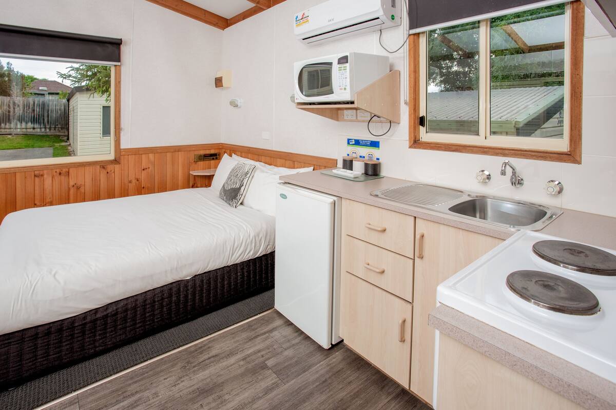 A compact kitchenette with a microwave, sink, and stove, a small bed with a pillow, and a window overlooking a backyard.