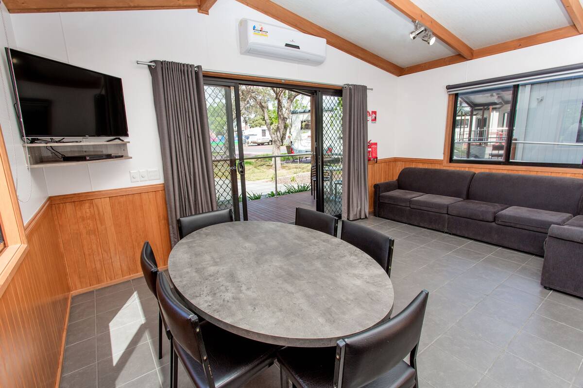 A room with a round table and chairs, a flat-screen TV mounted on the wall, a sofa, and a large window.