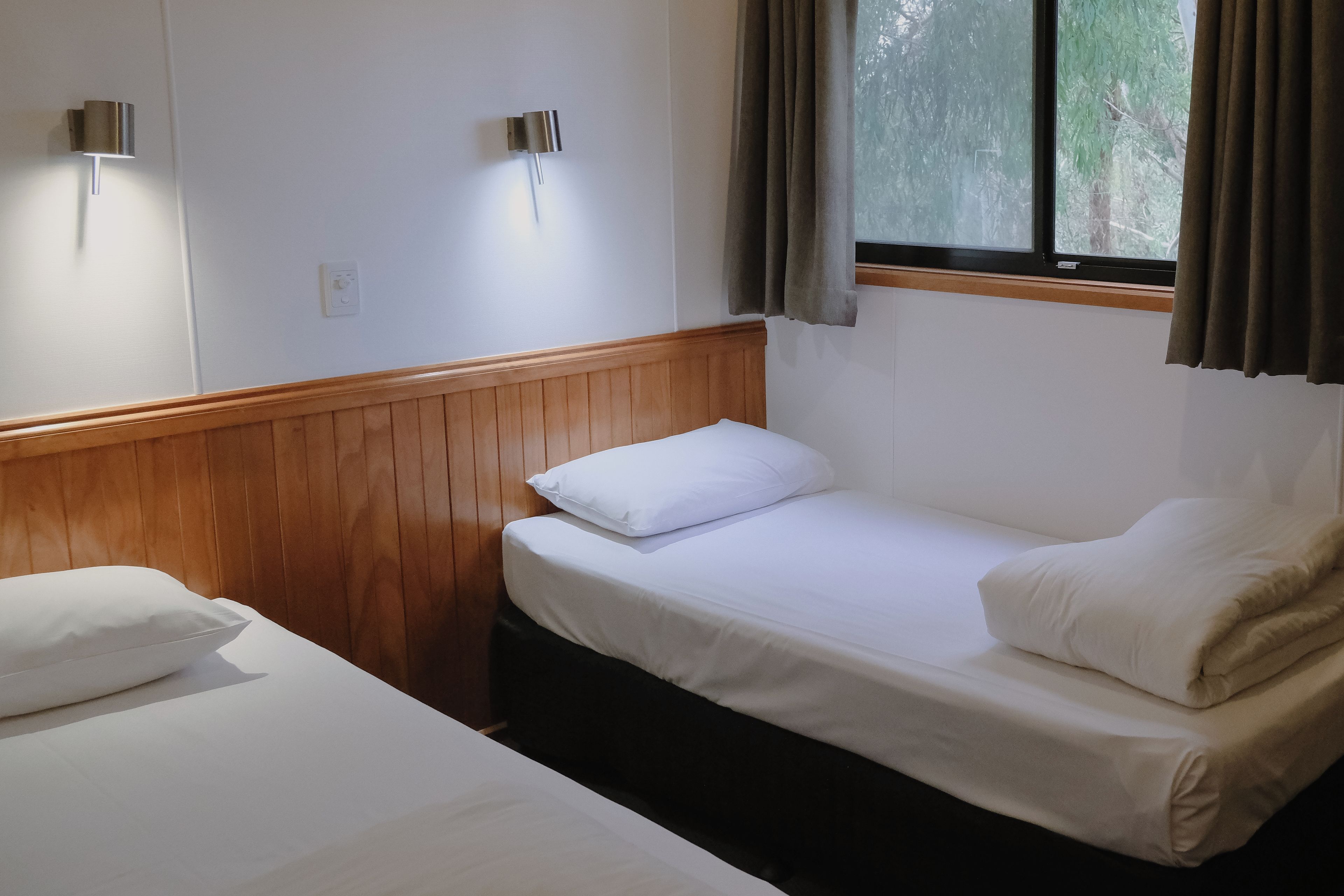 Habitación con dos camas individuales, cabecero de madera, ventana con cortinas e iluminación empotrada en la pared.