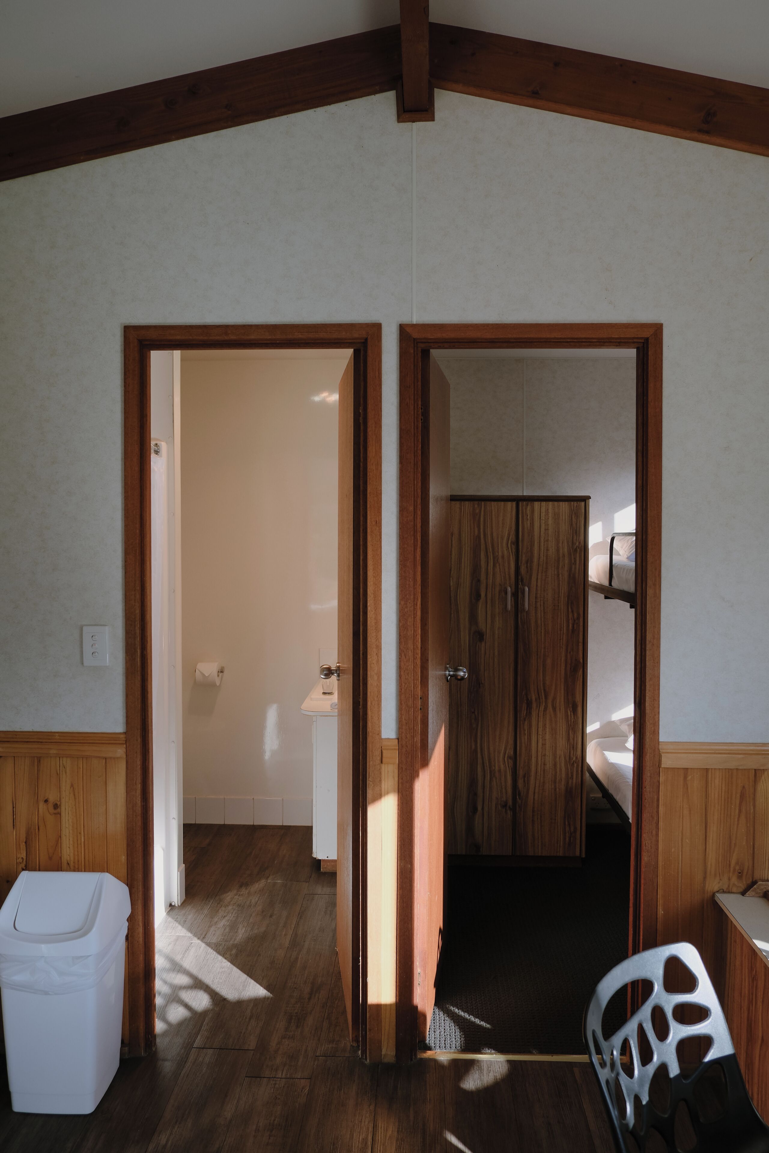 A room with wooden beams on the ceiling, a white door, a white trash bin, and a chair with a unique design.