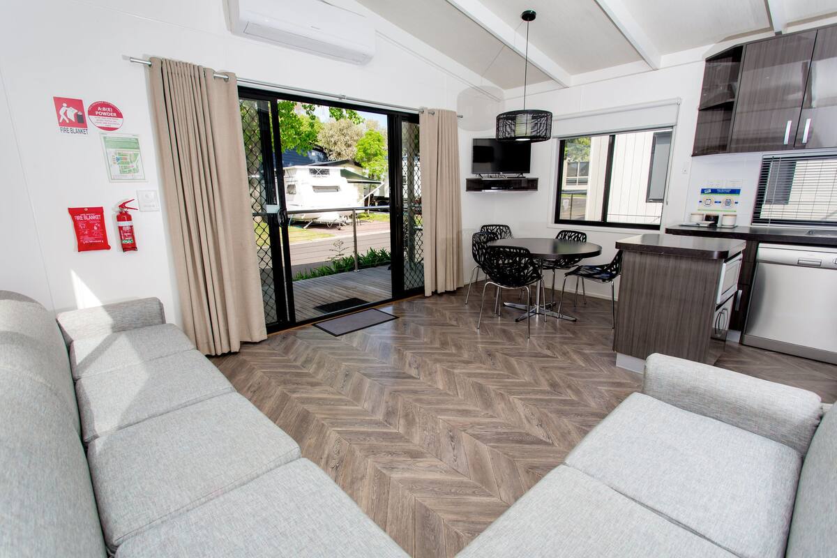 A modern, well-lit room with a herringbone wood floor, a sofa, a dining table with chairs, a kitchen area, and a sliding glass door leading to an outdoor area.