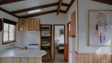 Cozinha compacta com armários de madeira, bancada branca e fogão. Há um corredor que leva a um quarto com cama e janela.