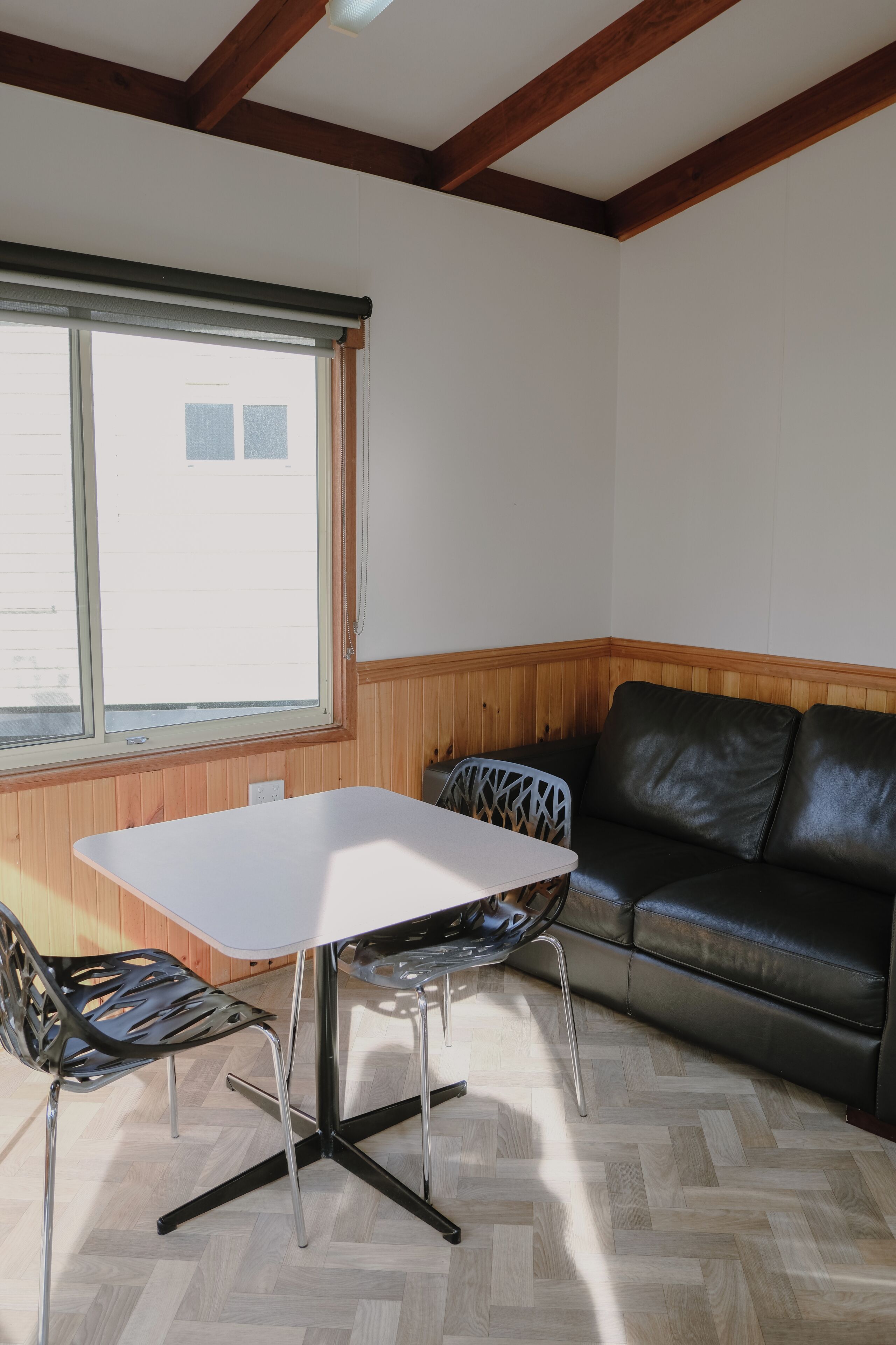 A room with a table and chairs, a window, a wooden wall, and a black leather sofa.