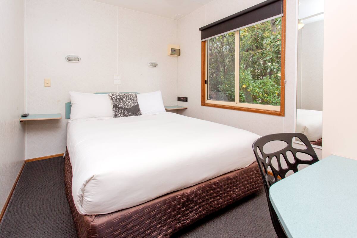A hotel room with a bed, a window with a view of trees, a small table, and a chair.