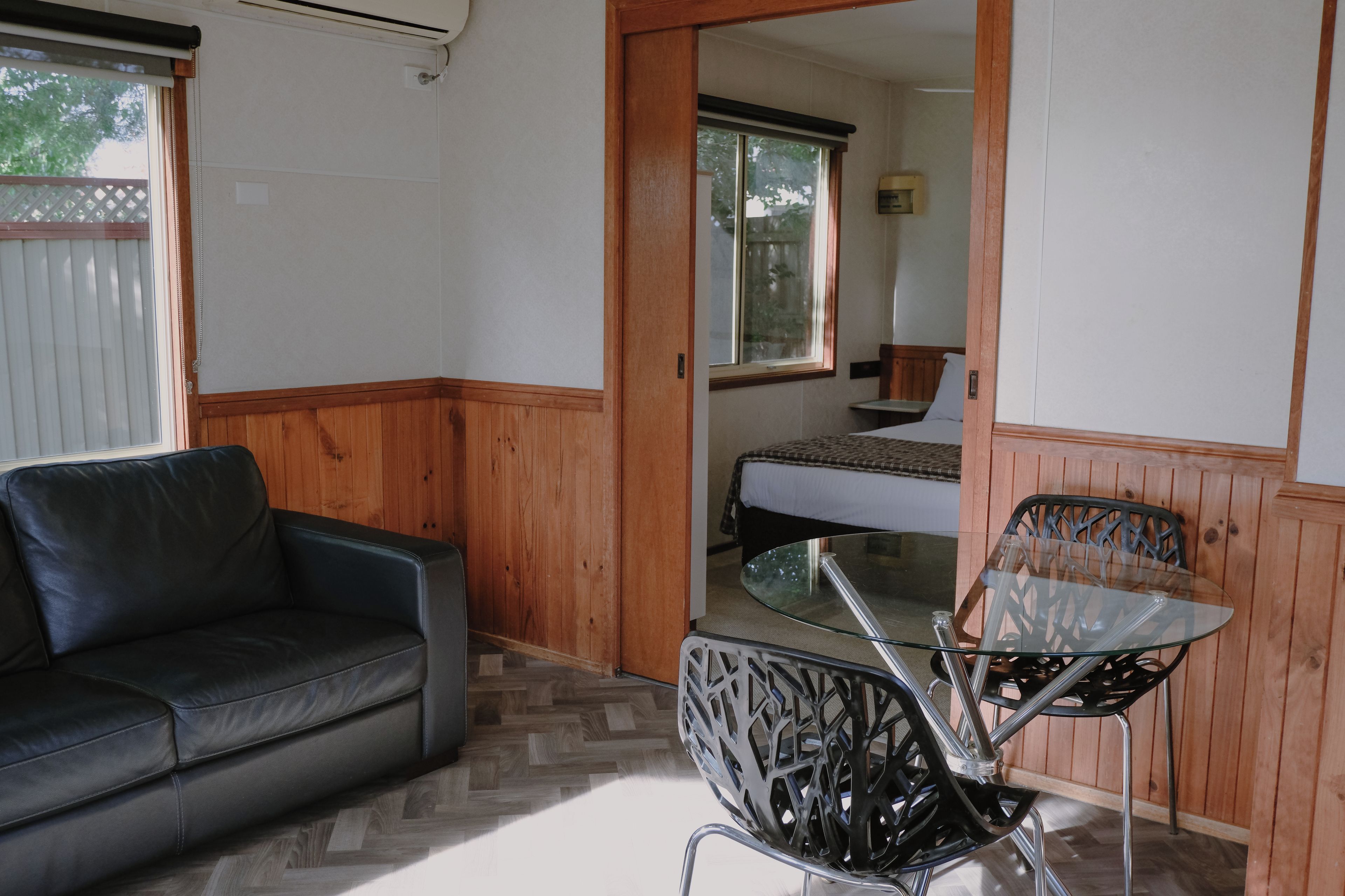 Quarto com sofá de couro preto, mesa de vidro com cadeiras de metal, piso de madeira, cama com colchão branco e janela com vista para árvores.