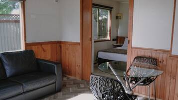 Quarto com sofá de couro preto, mesa de vidro com cadeiras de metal, piso de madeira, cama com colchão branco e janela com vista para árvores.