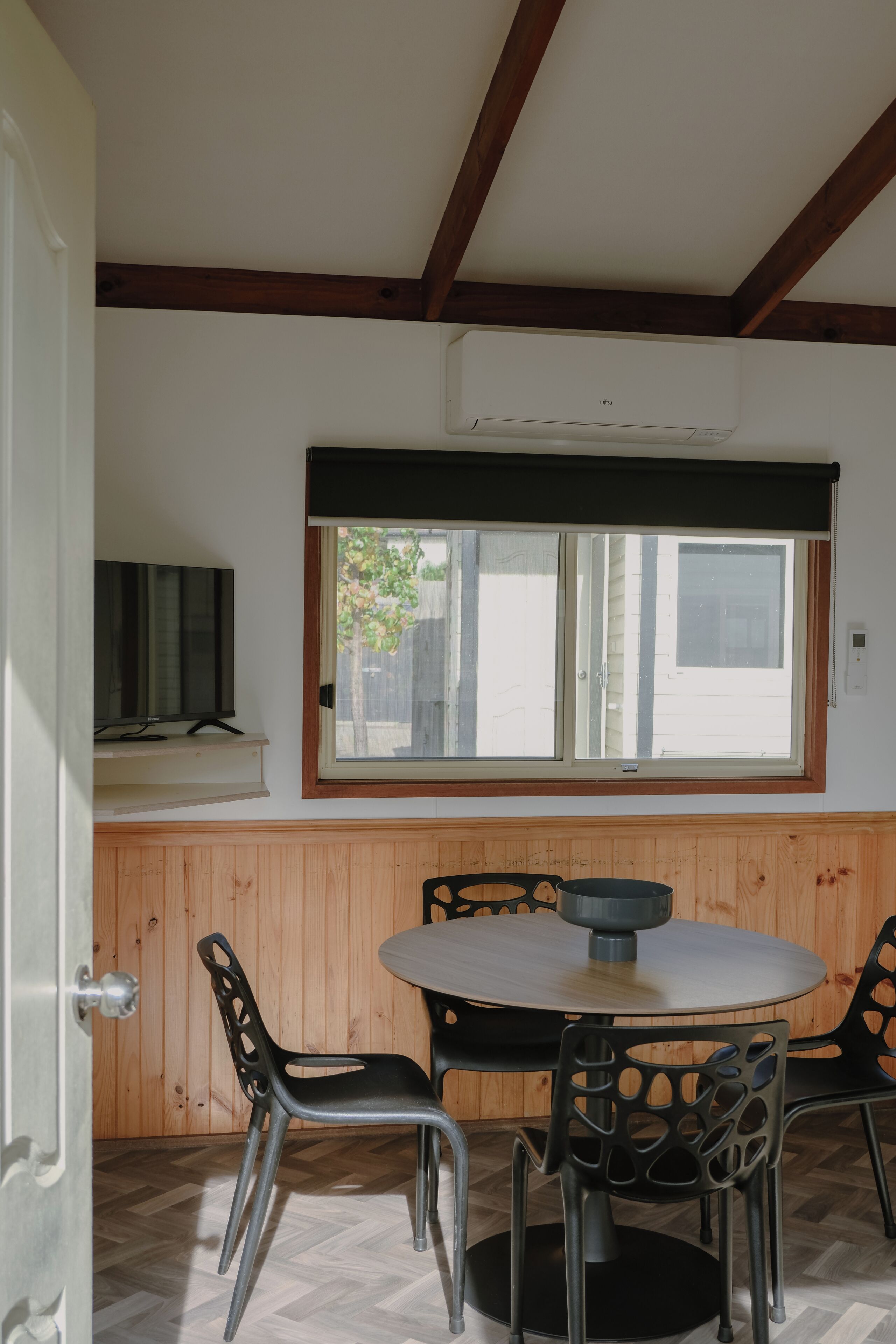 A room with a round table and chairs, a window with blinds, and a television on a shelf.