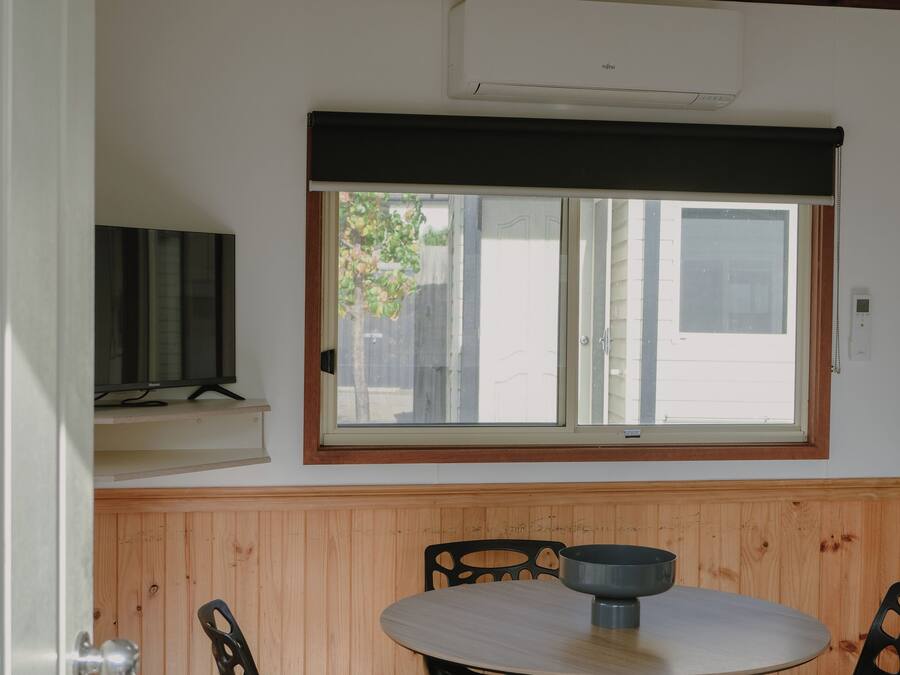 A room with a round table and chairs, a window with blinds, and a television on a shelf.