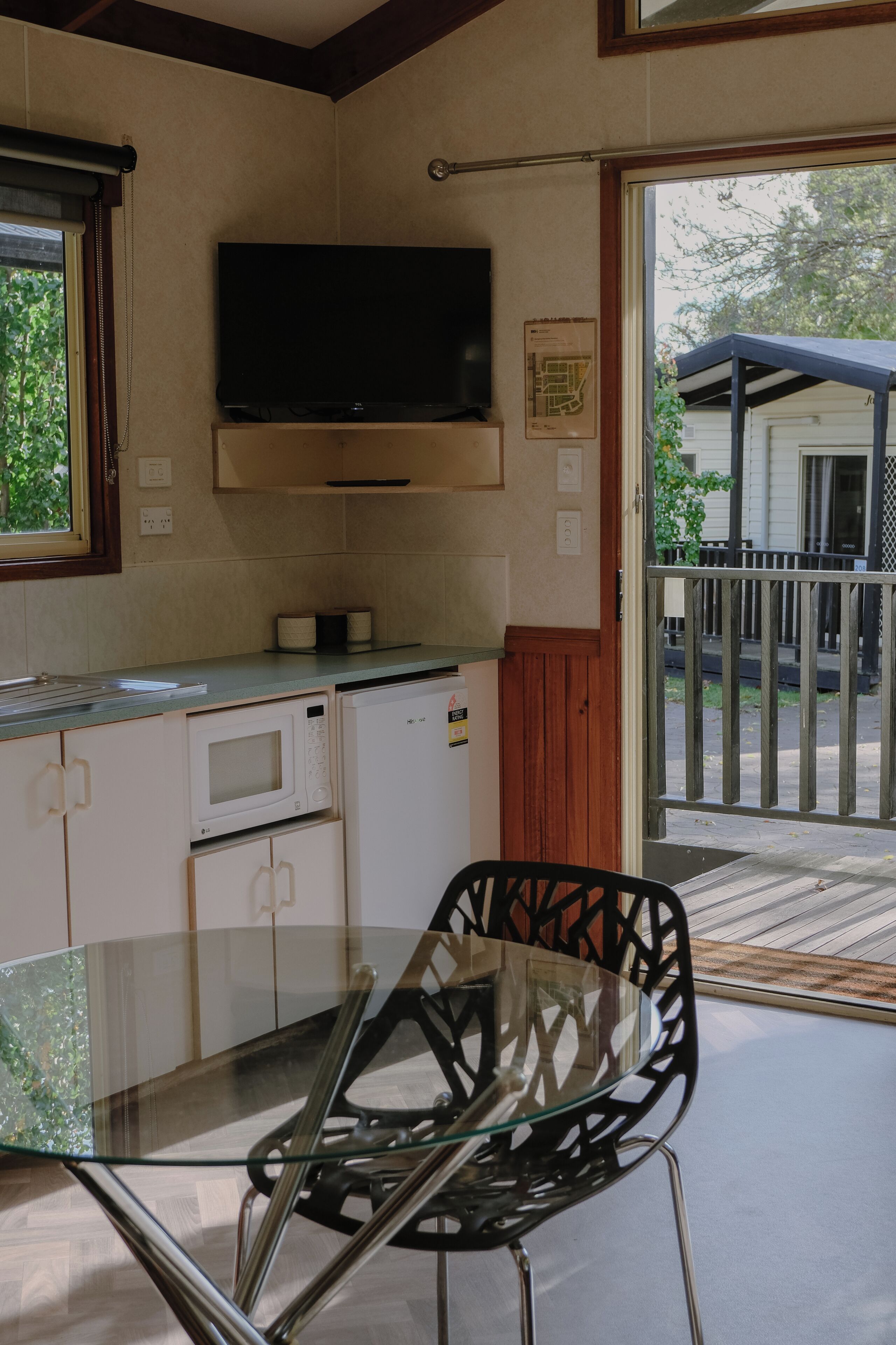 A kitchen with a flat-screen TV, a microwave, and a refrigerator. There is a glass table with a chair in front.