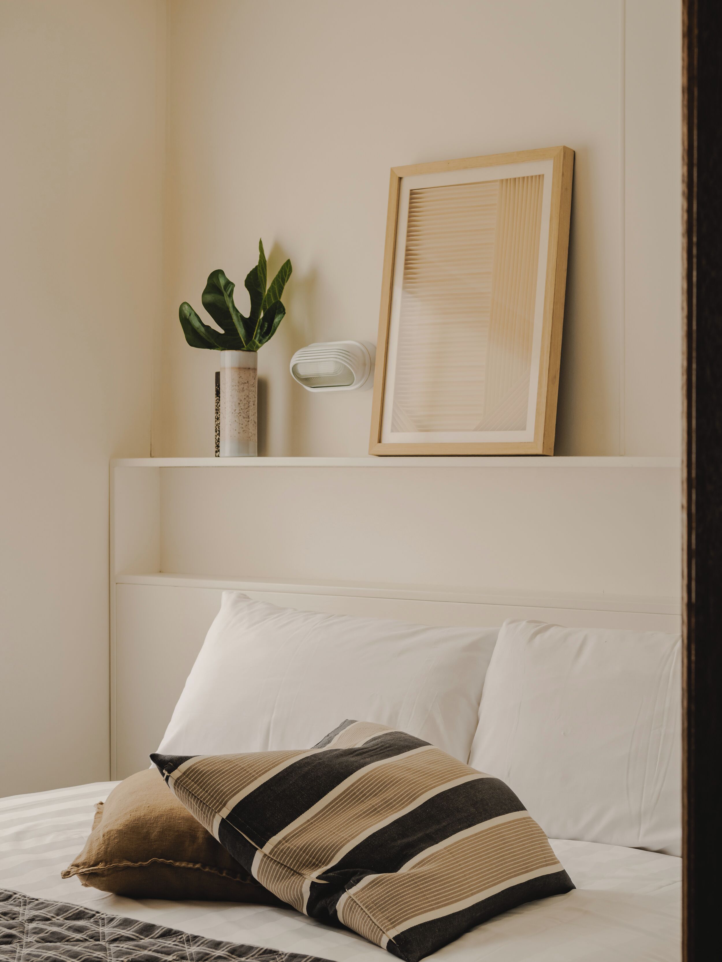 A bed with white bedding and striped pillows, a potted plant, and a framed picture on a bedside table.