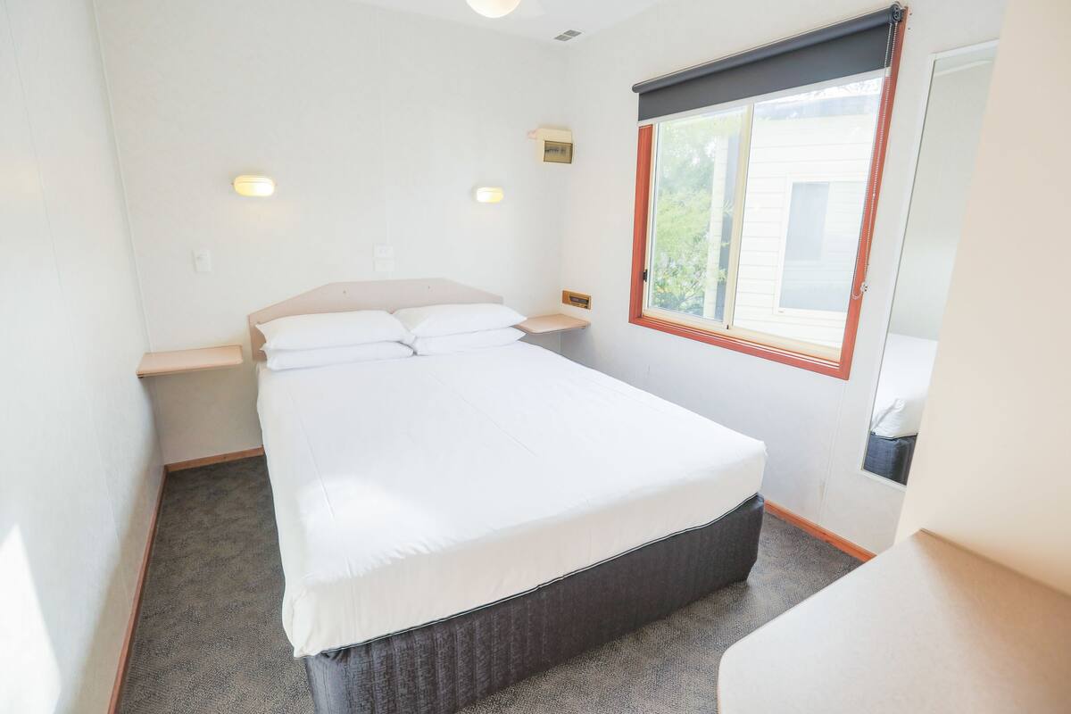 A single-bed room with a wooden headboard, a bedside table, a window with blinds, and a carpeted floor.