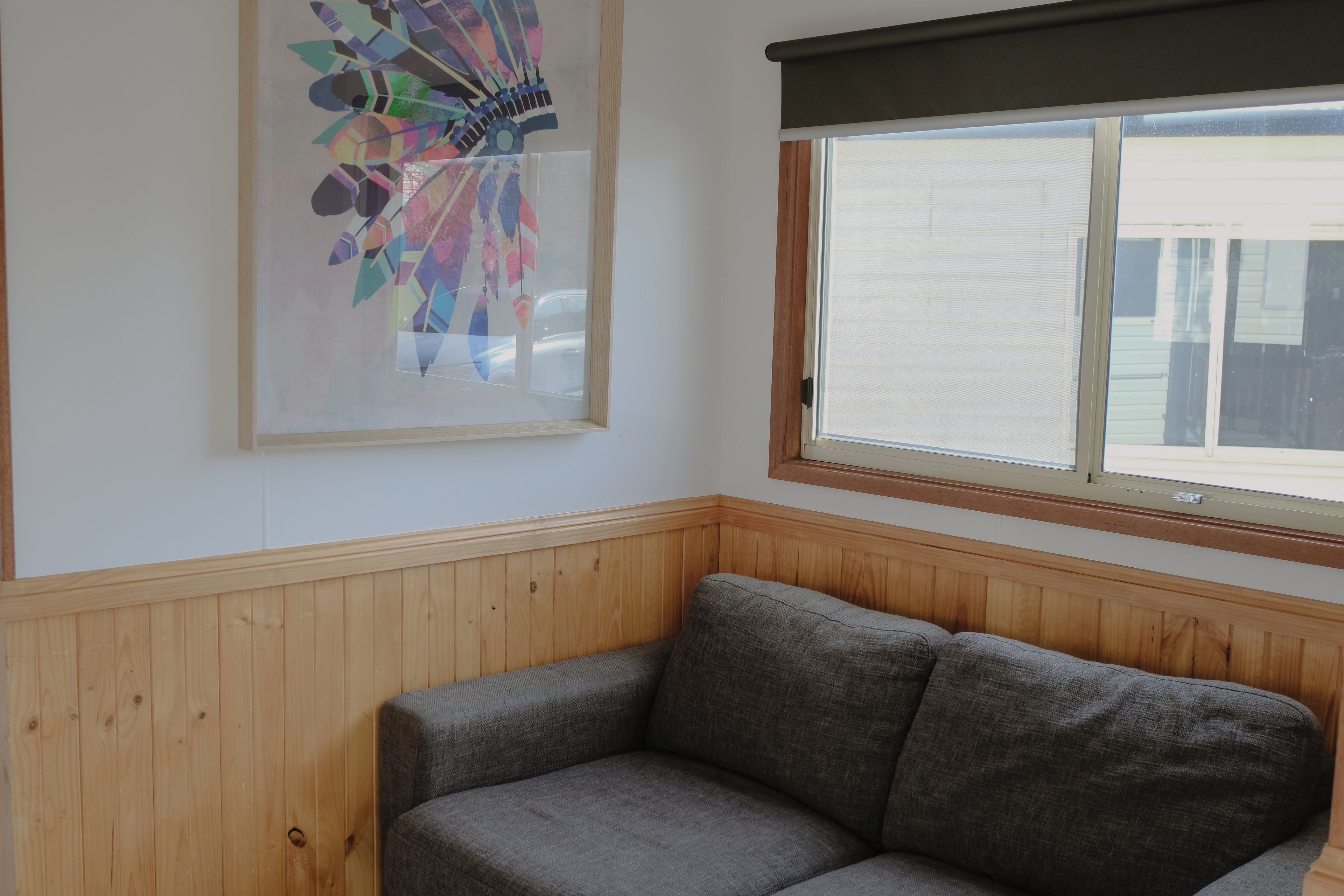 A corner room with a grey sofa, a wooden paneled wall, a window with blinds, and a colorful abstract painting.