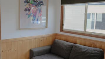 A corner room with a grey sofa, a wooden paneled wall, a window with blinds, and a colorful abstract painting.