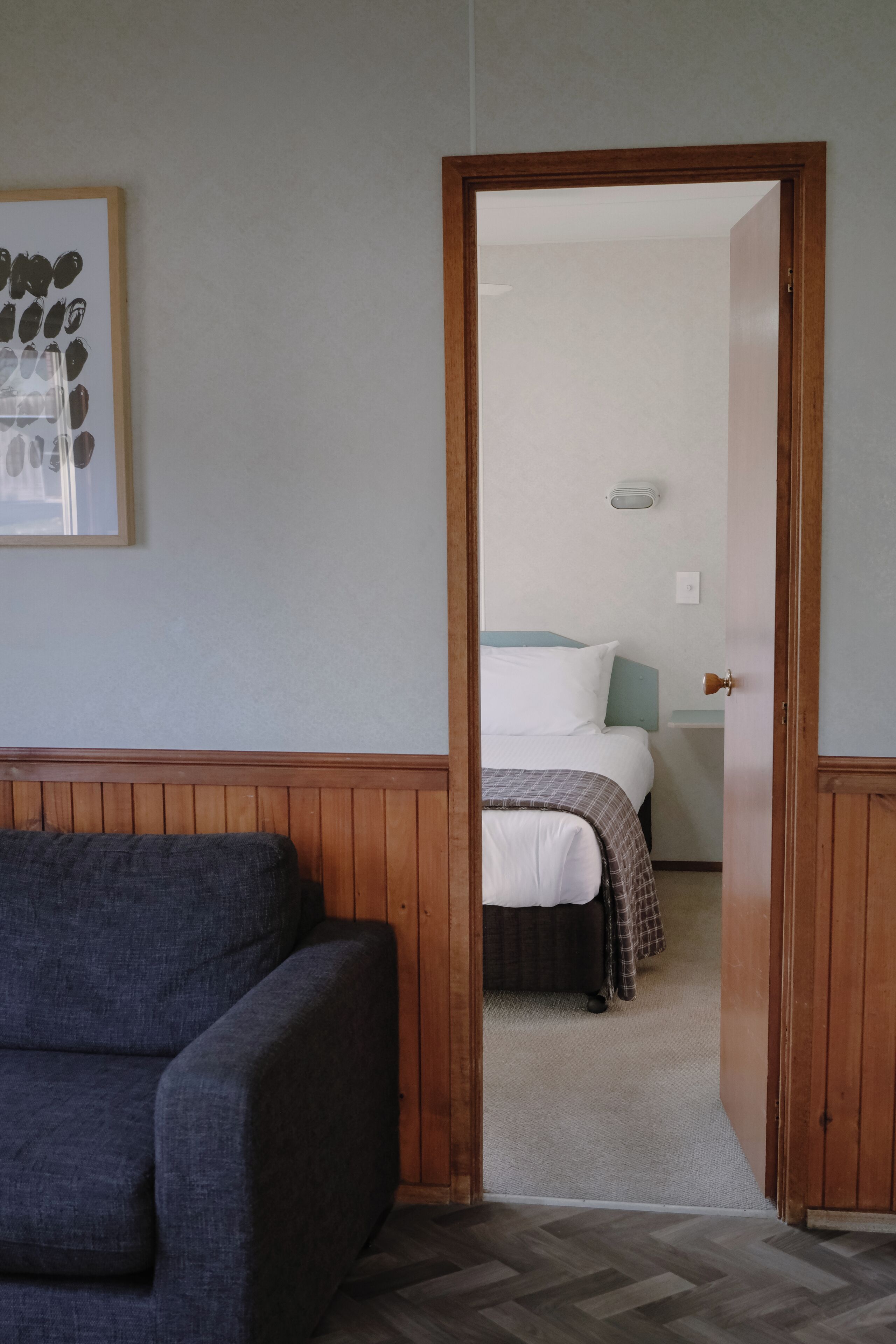 Una habitación con una puerta de madera que da a un dormitorio con una cama, una mesita de noche y una lámpara fijada a la pared.