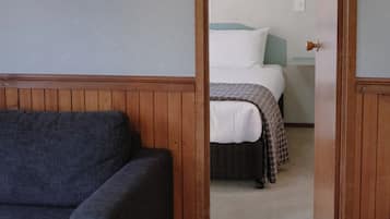 Una habitación con una puerta de madera que da a un dormitorio con una cama, una mesita de noche y una lámpara fijada a la pared.