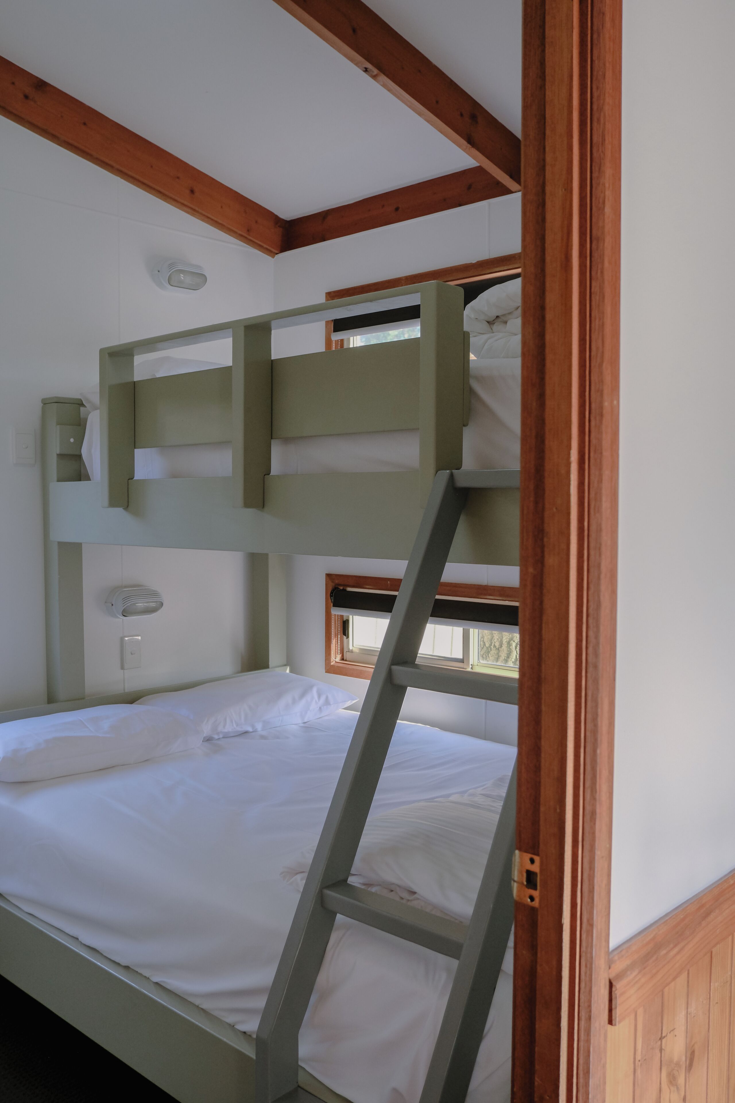 A bunk bed with a ladder, white bedding, a small window, and wooden beams.