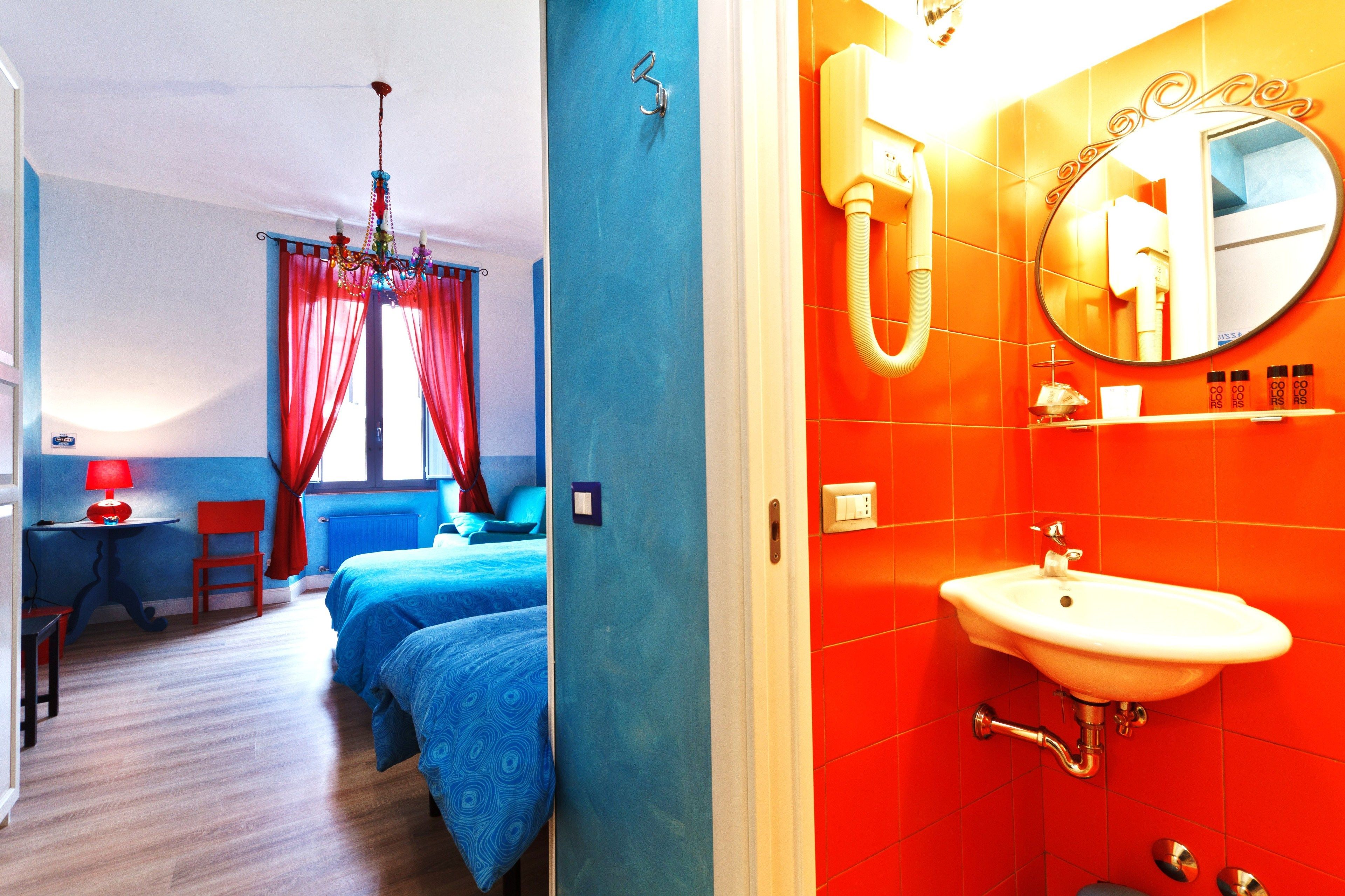 Une chambre d’hôtel aux couleurs bleu et rouge, avec un lit, un bureau doté d’une lampe et une salle de bain comprenant un lavabo et un miroir.