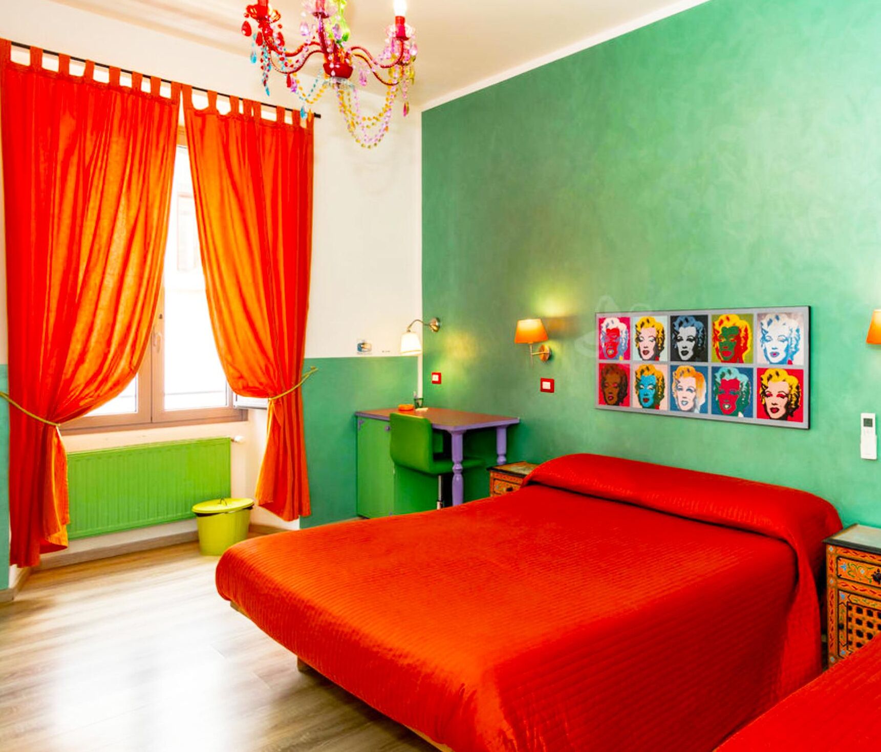 A room with a large bed, a desk, and a chair. There is a window with red curtains and a chandelier.