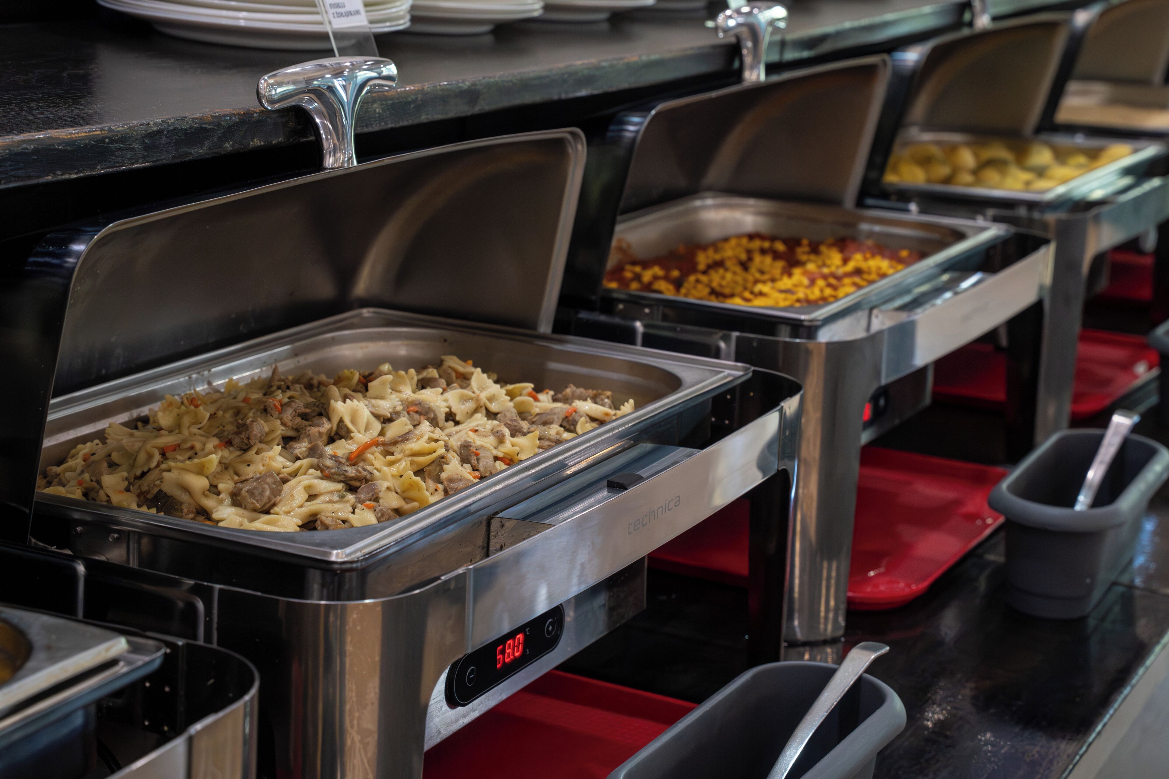 Un bufé con diversas opciones de comida en recipientes de acero inoxidable, un reloj digital y una bandeja roja.