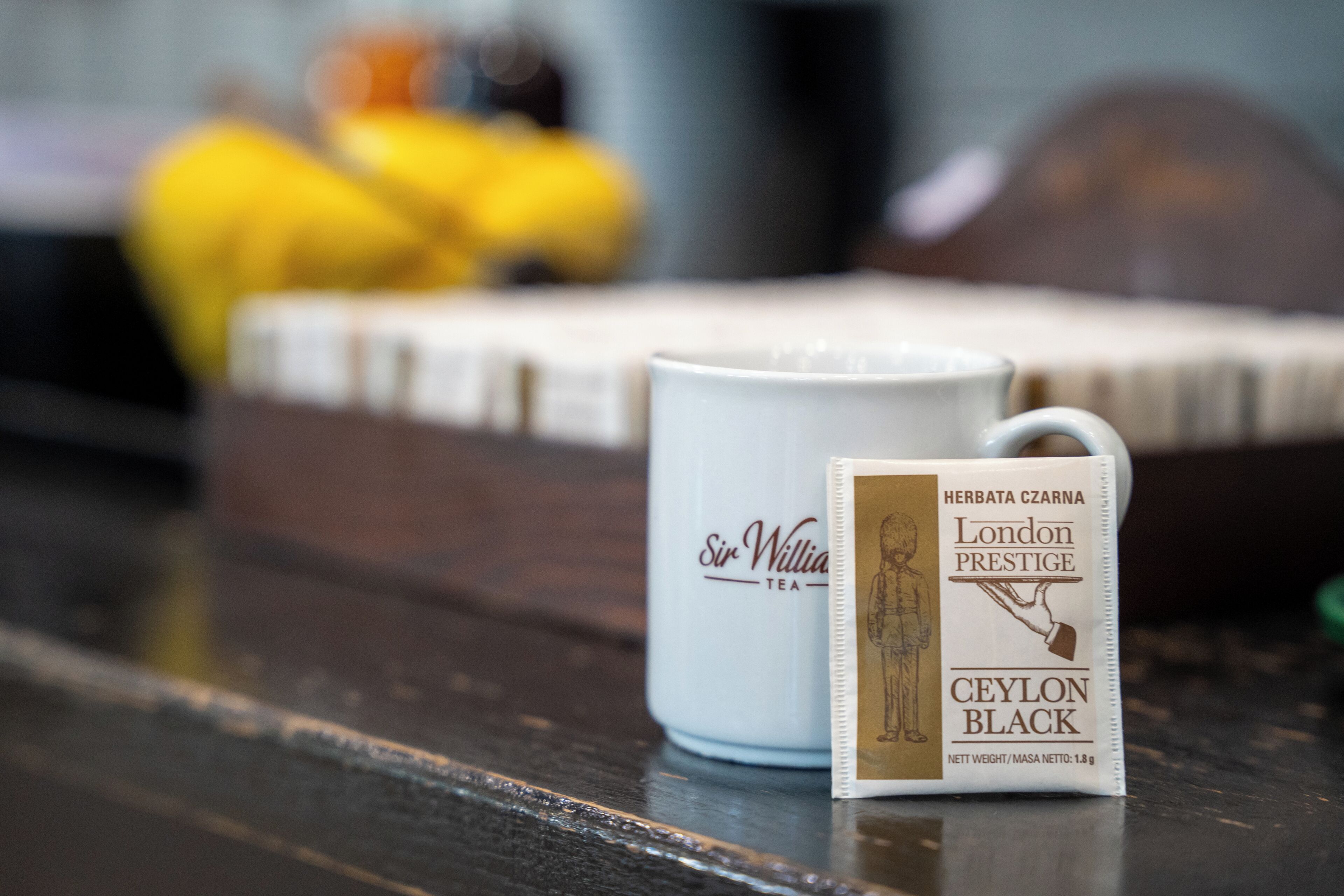 Una tazza bianca con l'etichetta 'Sir William Tea' e una confezione di tè etichettata 'Herbata Czarna London Prestige Ceylon Black' su un piano di legno.
