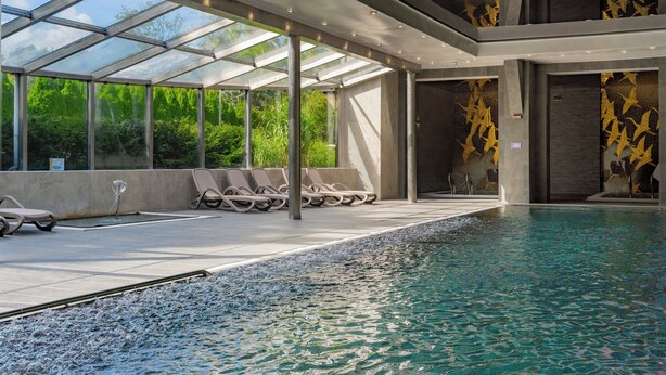 A modern indoor pool with clear glass walls, lounge chairs, and a stone wall with golden patterns. - Graal Spa (Swinoujscie)