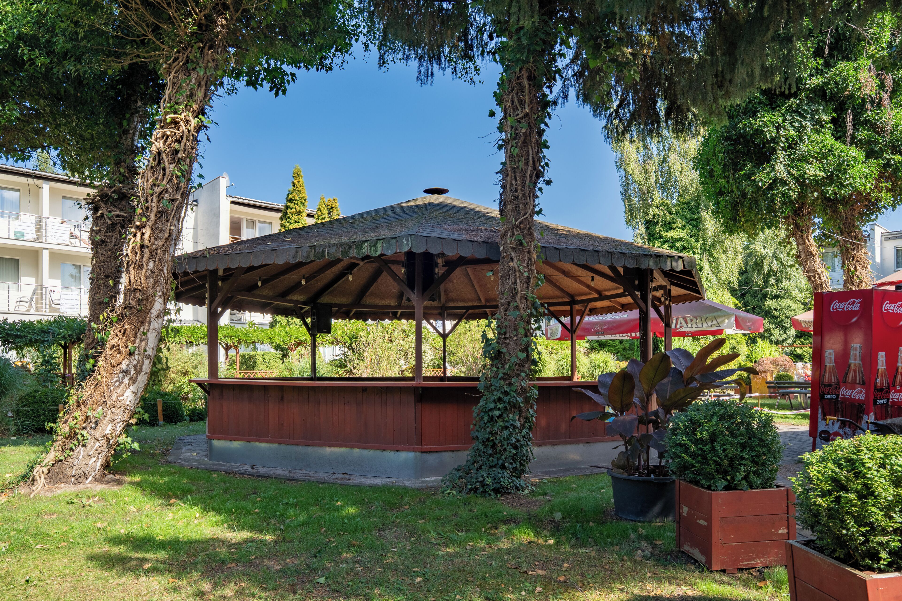 A covered pavilion with a roof, surrounded by trees and greenery, in a park-like setting.