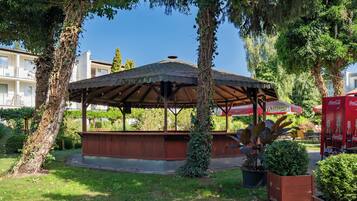 A covered pavilion with a roof, surrounded by trees and greenery, in a park-like setting.