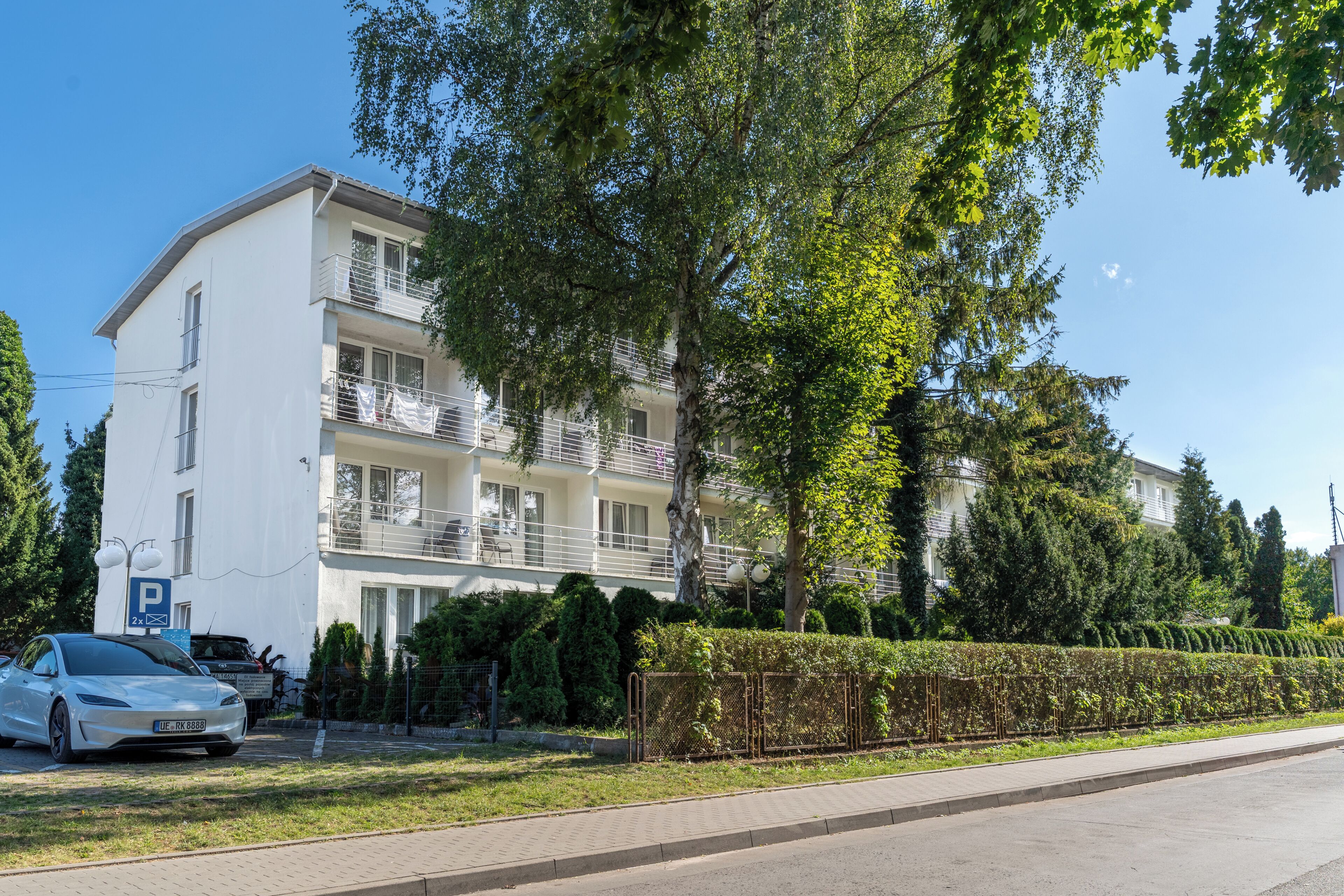 Un immeuble résidentiel à plusieurs étages, doté de balcons, d’un panneau indiquant un espace de stationnement et d’une haie le long du trottoir.
