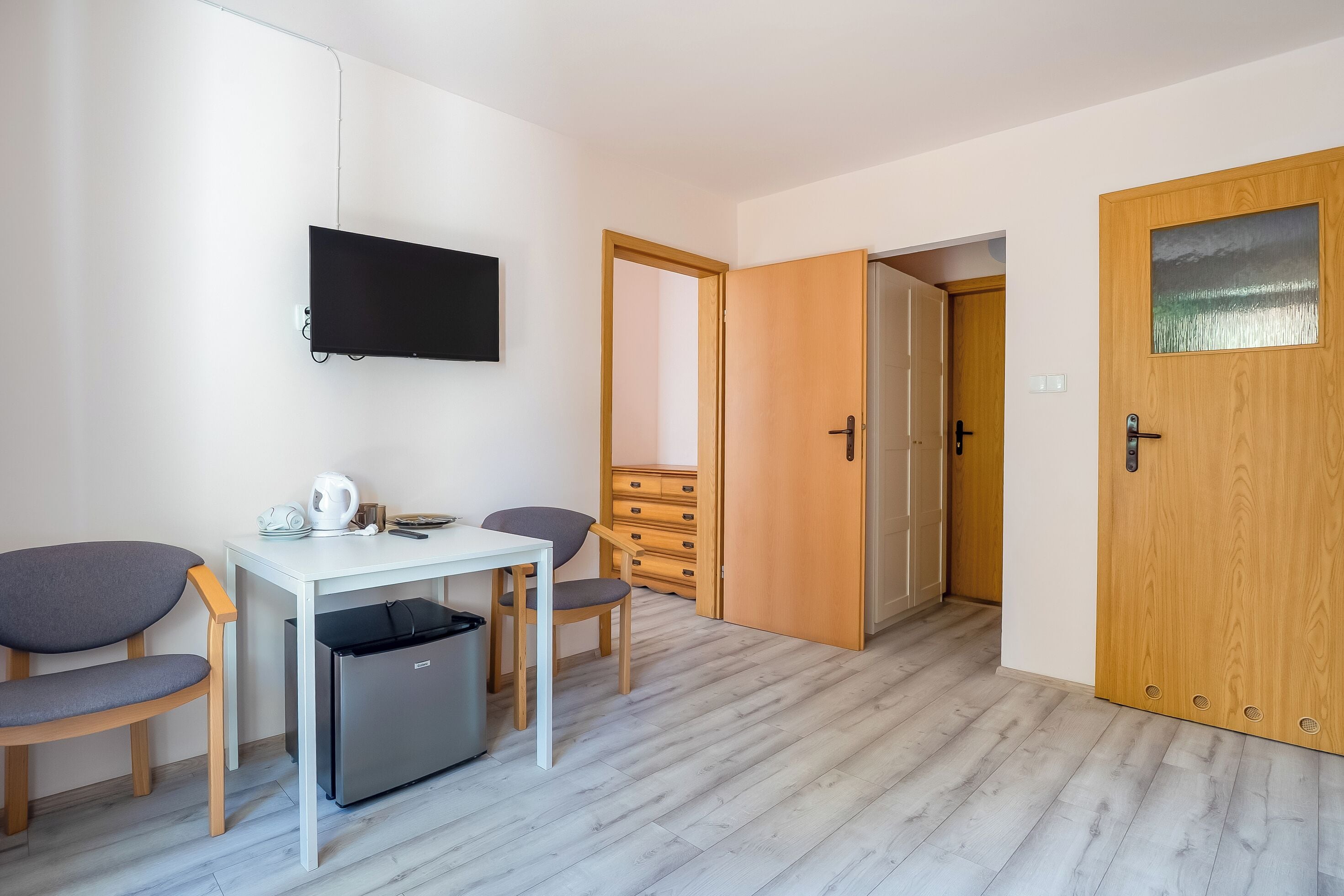 A room with a white desk, two chairs, a TV, a small fridge, and a wooden door with a glass panel.