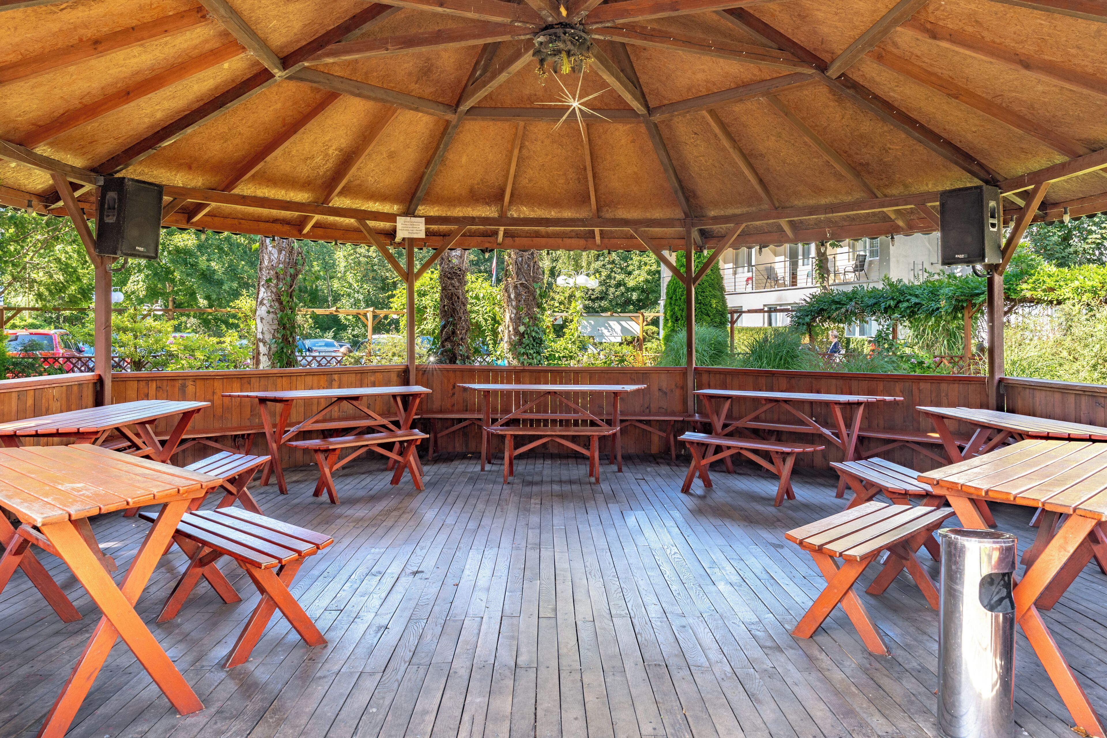 A covered outdoor area with wooden picnic tables and benches, surrounded by greenery.