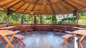 A covered outdoor area with wooden picnic tables and benches, surrounded by greenery.