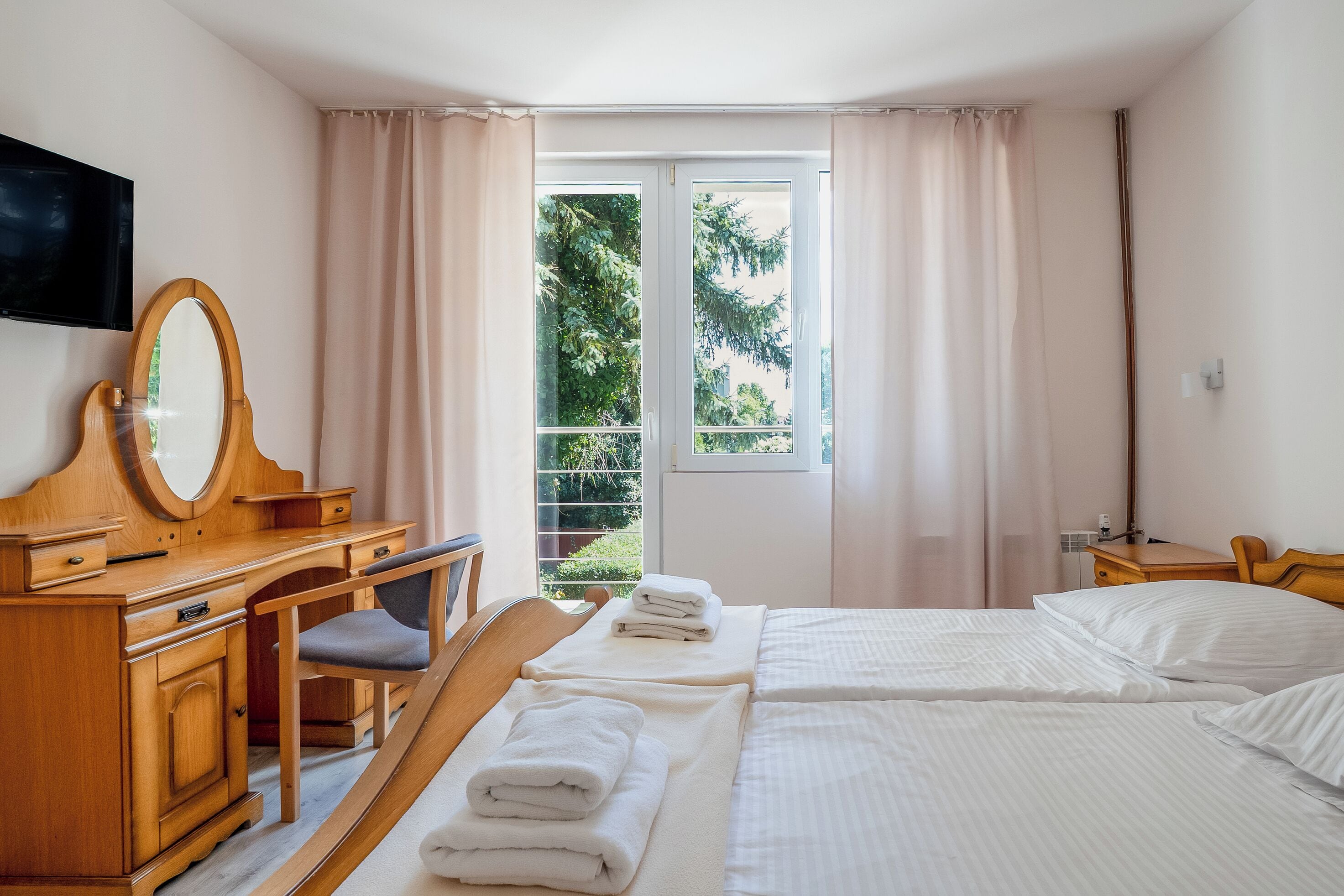 Ein Hotelzimmer mit einem Holztisch, einem Sessel, einem Bett mit weißer Bettwäsche, einem an der Wand befestigten Fernseher und einem Fenster mit Vorhängen.