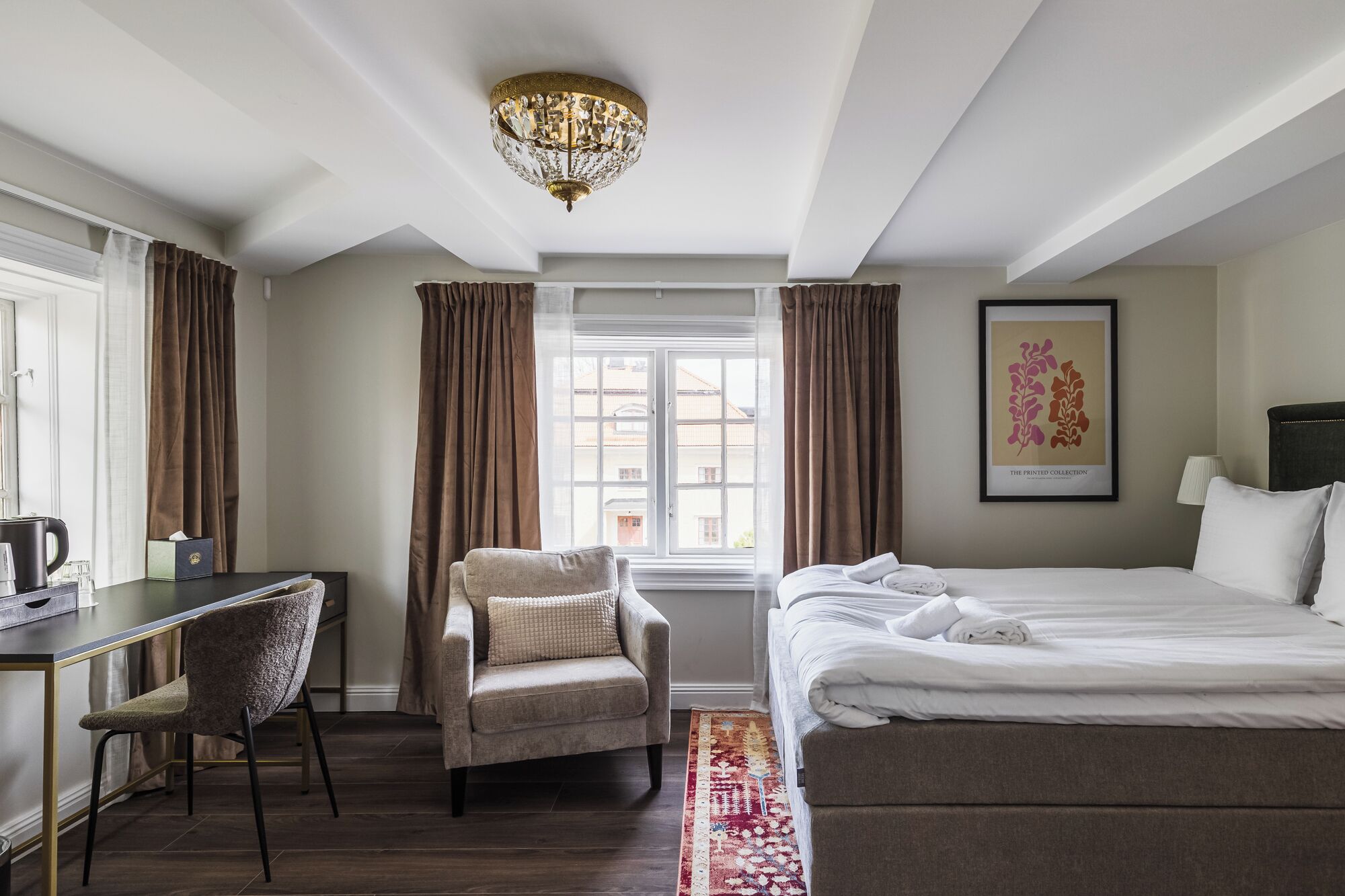 A hotel room with a bed, a desk, a chair, a chandelier, a window with curtains, and a framed picture on the wall.