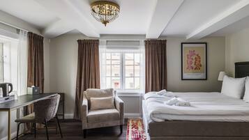 A hotel room with a bed, a desk, a chair, a chandelier, a window with curtains, and a framed picture on the wall.