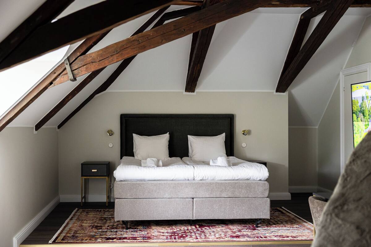 Un dormitorio con cama, mesita de noche, alfombra estampada y vigas de madera a la vista.