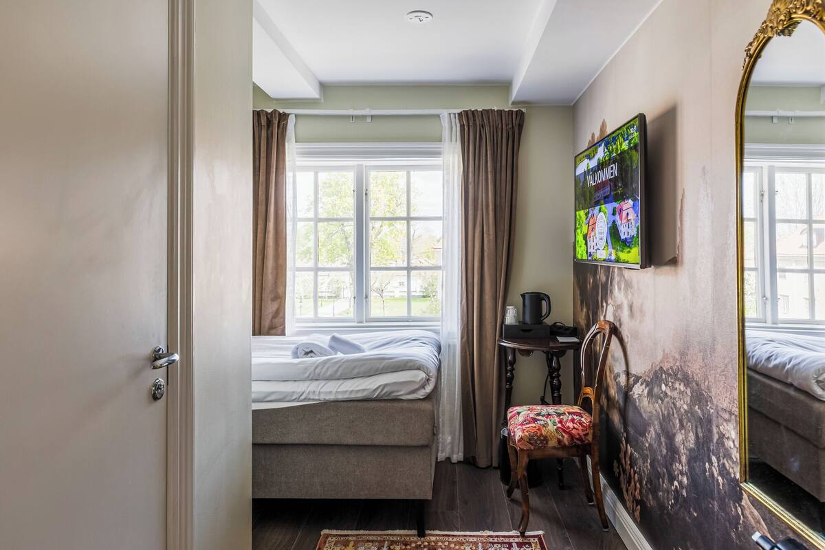 Una habitación con cama, una silla, una mesa con tetera, un espejo y un televisor montado en la pared.