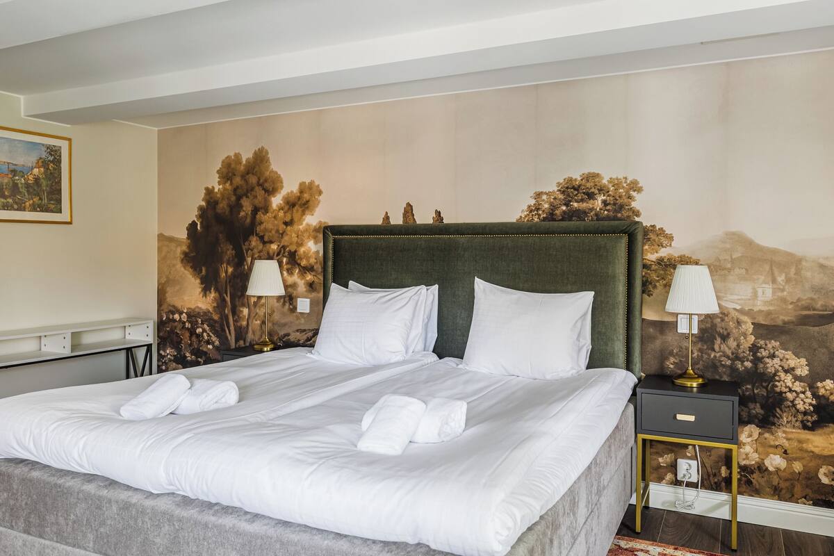 Una cama bien tendida con sábanas y toallas blancas, un cabecero tapizado en verde y un mural con paisaje en la pared.